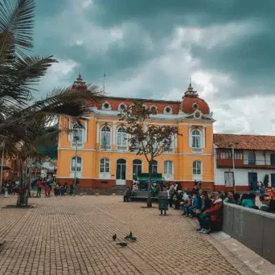 Plaza principal en Zipaquirá con un prominente edificio colonial amarillo de techo rojo y cúpulas. Palmeras frondosas y arquitectura tradicional flanquean la plaza, donde personas pasean y se relajan bajo un cielo nublado.