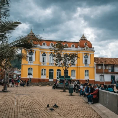 Plaza principal en Zipaquirá con un prominente edificio colonial amarillo de techo rojo y cúpulas. Palmeras frondosas y arquitectura tradicional flanquean la plaza, donde personas pasean y se relajan bajo un cielo nublado.