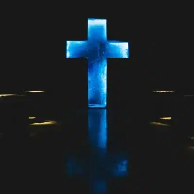 Cruz azul resplandeciente de material texturizado en un fondo oscuro, flanqueada por luces amarillas cálidas que se reflejan en el suelo.