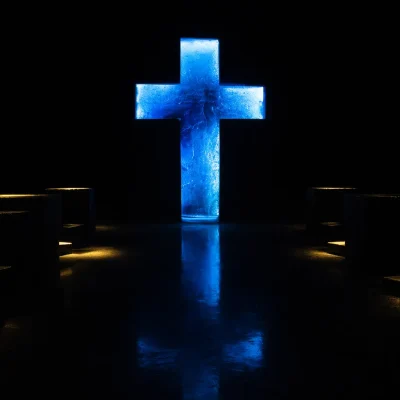 Cruz azul resplandeciente de material texturizado en un fondo oscuro, flanqueada por luces amarillas cálidas que se reflejan en el suelo.