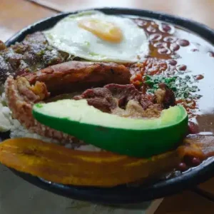 Primer plano de un plato abundante de comida tradicional (similar a la bandeja paisa) que contiene frijoles calientes, arroz blanco, carne asada, chorizo, chicharrón, huevo frito, plátano maduro frito y una rodaja de aguacate, servido sobre una mesa de madera.